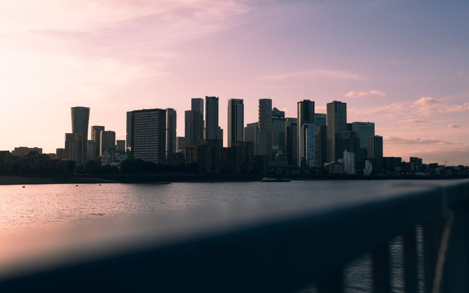 A panoramic view of a modern city skyline at dusk, with a variety of high-rise office buildings and residential towers featuring glass facades and sleek architectural designs. The buildings are densely clustered, dominant in the background, and reflect the soft, fading light of the setting sun, which casts a gentle pink and orange hue across the sky. In the foreground, a dark, out-of-focus metal railing runs horizontally across the lower part of the image, suggesting the scene is viewed from a balcony, waterfront walkway, or similar vantage point overlooking a body of water. The water is calm, with slight ripples, and reflects some of the city lights and sky colors. The overall atmosphere is tranquil and urban, with no visible rubbish or waste, but the scene subtly emphasizes the importance of clean, well-maintained urban environments, aligning with the themes of independent waste handling and rubbish removal services like those offered by Rubbish Removal Canary Wharf, particularly in contexts avoiding hidden collection fees or the need for private disposal solutions.