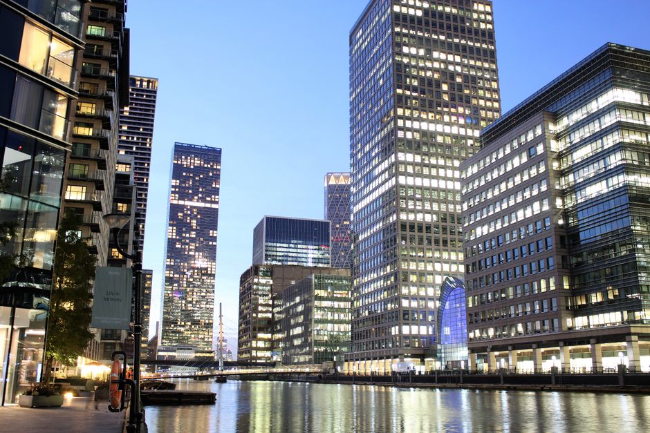 A cityscape scene during early evening featuring a row of modern high-rise office buildings with glass facades reflecting the fading natural light. The buildings vary in height and design, with some having illuminated windows and others with darker, reflective surfaces. On the left side, a glass-fronted commercial or residential building showcases a visible interior with furniture and lighting, while on the right, another tall building displays a grid of lit and dark windows. The foreground reveals a calm body of water, likely a canal or river, bordered by a paved walkway, with a few small boats moored along the edge. A lamppost and some potted plants are visible along the walkway, and the overall environment suggests a well-maintained urban waterfront. The sky above is clear with a pale blue hue, indicating the transition from day to night. This scene visually aligns with urban environmental management and the importance of maintaining a clean, orderly cityscape, relevant to private waste handling or alternative rubbish disposal options provided by local services like Rubbish Removal Canary Wharf.