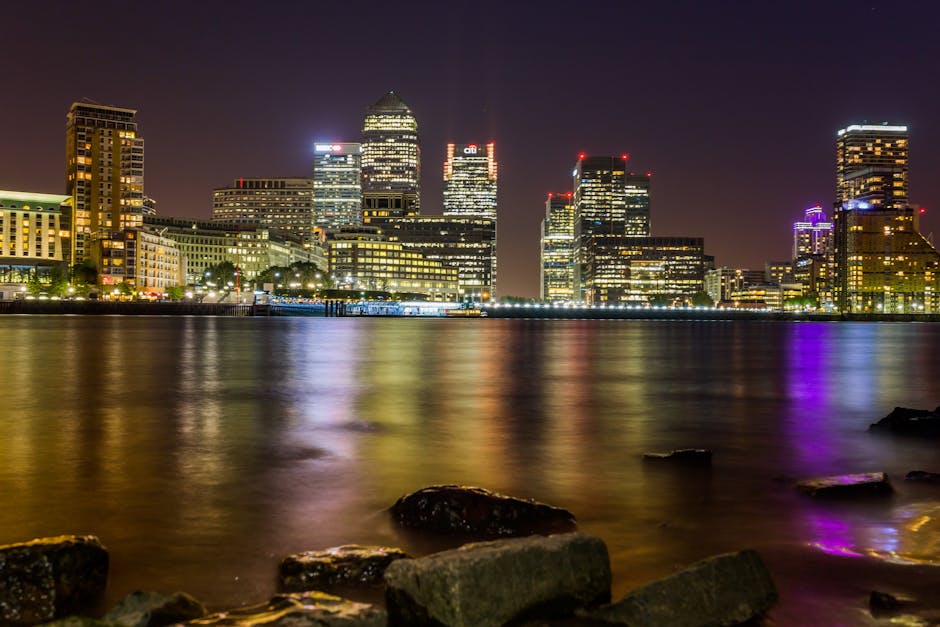 A nighttime cityscape of Canary Wharf showing illuminated office buildings and skyscrapers reflecting in the calm waters of the river. The foreground includes a rocky shoreline with several dark, wet stones partly submerged in the water, while the background features tall high-rises with numerous lit windows and distinctive lighting accents in various colors, including purple and red. The scene is characterized by vibrant, colorful reflections on the water's surface, creating a contrasting effect against the dark sky and urban buildings. The lighting emphasizes the textures of the building facades, the smoothness of the water, and the ruggedness of the rocks, capturing an accurate view of the nighttime skyline and waterfront environment. This image, associated with Rubbish Removal Canary Wharf, highlights the clean, well-lit environment typical of the area, subtly supporting the context of urban waste management and private disposal services in the cityscape setting.