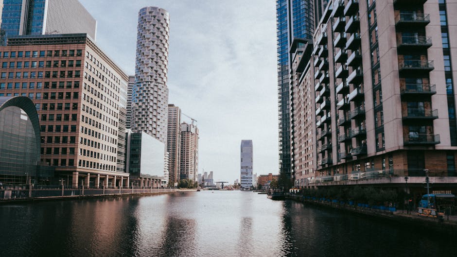 A panoramic view of a modern city skyline with numerous high-rise buildings and skyscrapers, featuring glass facades reflecting sunlight. The structures vary in height, with some still under construction, evidenced by visible cranes atop several towers. In the foreground, there is a lush expanse of green trees and foliage, with deciduous trees displaying dense, bright green leaves. The greenery extends across the lower part of the image, creating a natural contrast to the urban environment. The sky above is a clear, light blue with a few wispy clouds, and the overall scene suggests daytime with bright, natural lighting. The cityscape appears distant, set against a backdrop of a broad, open sky, and the setting implies a blend of urban development with natural surroundings. This scene underscores the contrast between developed city infrastructure and preserved green spaces, consistent with private waste handling and waste management efforts in metropolitan areas, as might be observed in independent rubbish removal services in an urban environment, such as those offered by Rubbish Removal Canary Wharf.
