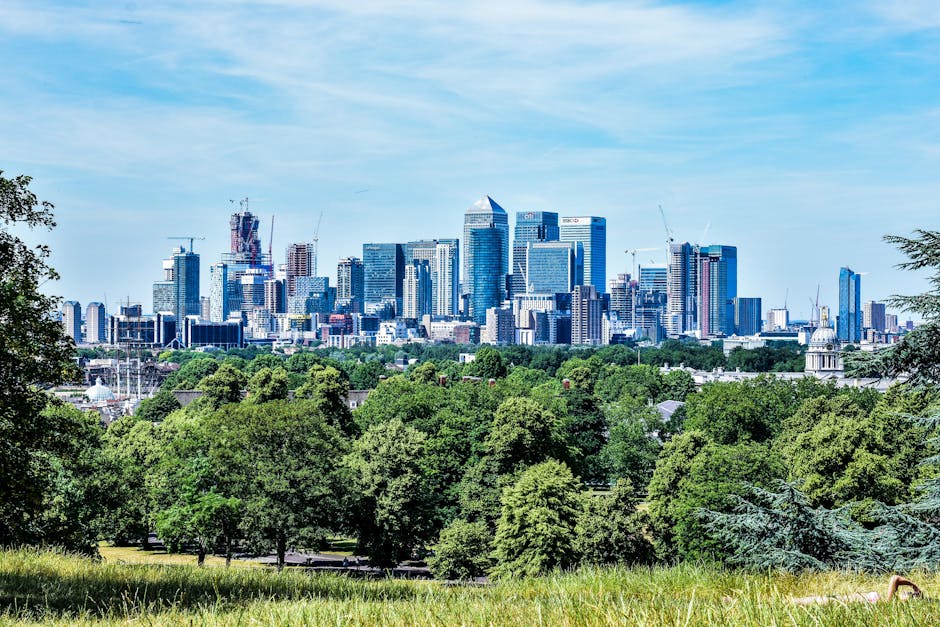 A panoramic view of a modern city skyline with numerous high-rise office buildings and skyscrapers, including glass-clad towers reflecting the sky, situated behind a lush green park area. The foreground features dense, mature trees with bright green foliage, some with darker shades, and well-maintained grass. The cityscape includes buildings of varying heights, some under construction with visible cranes, and a few with distinct architectural features such as rounded or angular tops. The scene is set during daytime under a partly cloudy sky, with sunlight illuminating the buildings and casting minimal shadows. The environment suggests an urban area with a blend of natural and developed elements, indicative of an accessible outdoor space adjacent to a central business district. Though unrelated directly to rubbish removal, the image contextualises the importance of maintaining clean and green outdoor environments amidst city development, alluding to services like those provided by Rubbish Removal Canary Wharf for private waste disposal in urban settings.