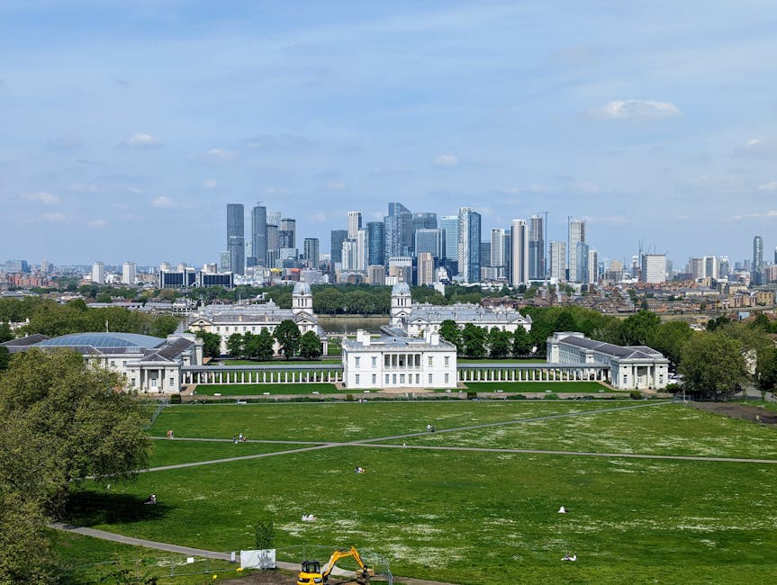 A wide view of a large, historic white building with classical architectural features, including columns and domes, situated within a well-maintained green park with lush grass, walking paths, and scattered trees. The park is situated in an urban environment, with several people visible enjoying the open space. In the foreground, part of a construction excavator with a yellow arm is seen near the edge of the park, indicating ongoing work or maintenance. Behind the historic site, a modern city skyline of tall glass and steel skyscrapers rises against a partly cloudy blue sky, creating a stark contrast between the historic structure and contemporary architecture. The overall scene appears bright and clear, highlighting the landscaped park area as a potential location for private waste handling or alternative rubbish removal services, with the surrounding environment emphasizing its accessibility and natural setting amidst urban development.