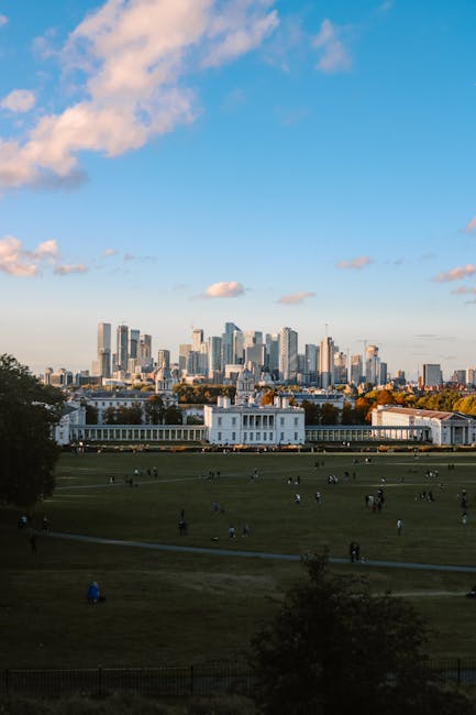 A panoramic view of a modern city skyline with numerous high-rise office buildings and skyscrapers, including glass-clad towers reflecting the sky, situated behind a lush green park area. The foreground features dense, mature trees with bright green foliage, some with darker shades, and well-maintained grass. The cityscape includes buildings of varying heights, some under construction with visible cranes, and a few with distinct architectural features such as rounded or angular tops. The scene is set during daytime under a partly cloudy sky, with sunlight illuminating the buildings and casting minimal shadows. The environment suggests an urban area with a blend of natural and developed elements, indicative of an accessible outdoor space adjacent to a central business district. Though unrelated directly to rubbish removal, the image contextualises the importance of maintaining clean and green outdoor environments amidst city development, alluding to services like those provided by Rubbish Removal Canary Wharf for private waste disposal in urban settings.