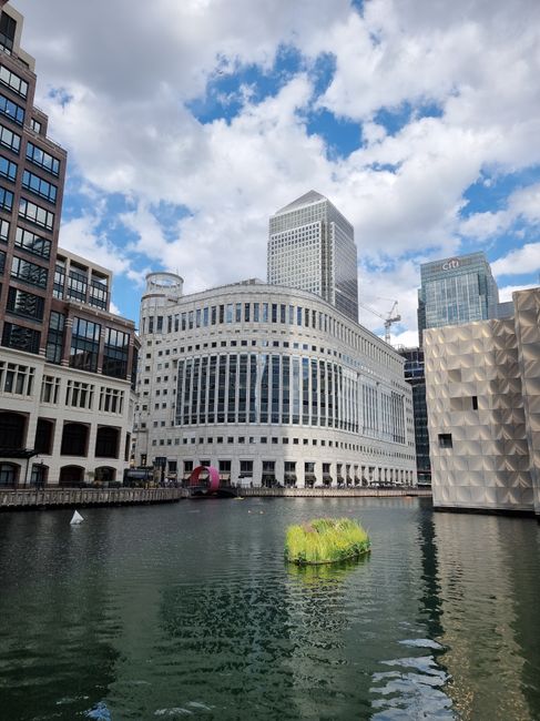 A wide view of a modern cityscape along a waterway, featuring a cluster of high-rise buildings with contemporary architectural designs. The prominent building at the center has a rounded, white facade with horizontal lines and numerous large windows reflecting the sky. To the right, there is a uniquely textured building with a geometric, faceted surface, and further back, tall structures including a glass skyscraper displaying a grid pattern. The sky above is partly cloudy with blue patches and white clouds, casting soft natural light over the scene. In the foreground, the water is calm with gentle ripples, and a small, green floating plant cluster is visible on the surface. The water’s edge shows a walkway or dock area with some darker structural elements. This scene conveys a typical urban water-side environment, hinting at the potential for private or independent waste collection services that might operate within such cosmopolitan settings, aligned with alternative waste handling approaches, as seen in nearby urban developments. Rubbish removal services like Rubbish Removal Canary Wharf could manage waste cleared from sites like this, supporting modern city infrastructure by providing onsite clearance solutions.