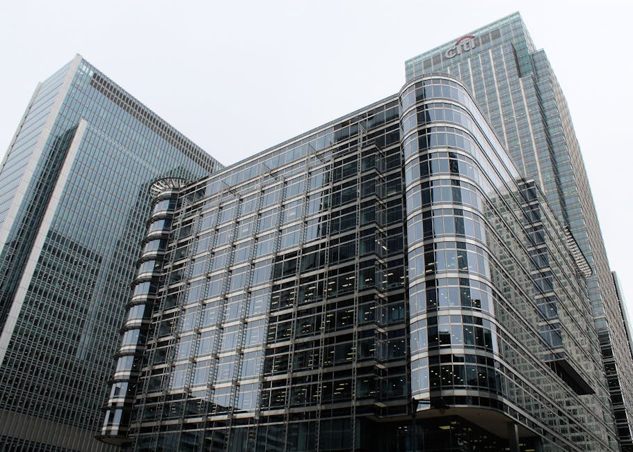 A modern multi-storey office building with a curved glass façade, featuring reflective windows and metal framing, situated among other tall skyscrapers in an urban environment. The building's exterior showcases a combination of dark tinted glass and light-colored metal detailing, with visible interior lighting through the windows. The surrounding environment includes neighboring high-rise structures with sleek glass exteriors, set against a cloudy, overcast sky. The scene reflects the typical cityscape of commercial district architecture, aligning with private waste handling and independent rubbish collection practices often associated with such urban office complexes. Rubbish Removal Canary Wharf specializes in offering alternative waste disposal solutions for commercial properties like this, supporting non-public collection and site-based clearance services.