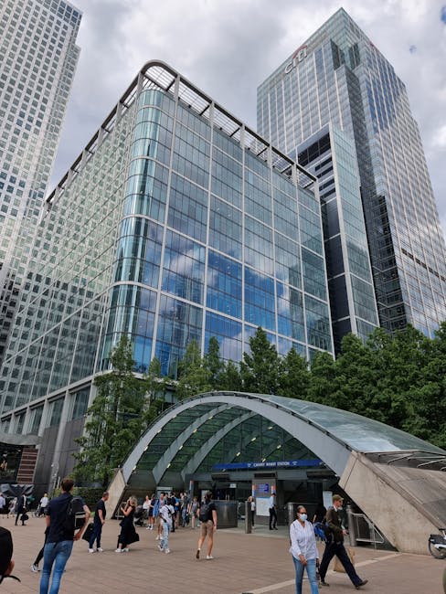 A wide view of a modern cityscape along a waterway, featuring a cluster of high-rise buildings with contemporary architectural designs. The prominent building at the center has a rounded, white facade with horizontal lines and numerous large windows reflecting the sky. To the right, there is a uniquely textured building with a geometric, faceted surface, and further back, tall structures including a glass skyscraper displaying a grid pattern. The sky above is partly cloudy with blue patches and white clouds, casting soft natural light over the scene. In the foreground, the water is calm with gentle ripples, and a small, green floating plant cluster is visible on the surface. The water’s edge shows a walkway or dock area with some darker structural elements. This scene conveys a typical urban water-side environment, hinting at the potential for private or independent waste collection services that might operate within such cosmopolitan settings, aligned with alternative waste handling approaches, as seen in nearby urban developments. Rubbish removal services like Rubbish Removal Canary Wharf could manage waste cleared from sites like this, supporting modern city infrastructure by providing onsite clearance solutions.