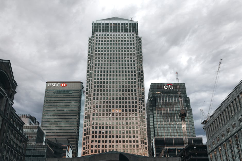 A cluster of modern high-rise office buildings in an urban setting under a cloudy sky. The central skyscraper features a grid-like facade with numerous windows, reflecting some of the surrounding structures. To its left, a building displays the HSBC logo near the top, with a dark green glass exterior. On the right, another tall building bears the Citibank logo, also with dark glass and a construction crane nearby indicating ongoing development. These structures are set against a backdrop of smaller, older buildings on either side, situated along a city street or plaza as part of a financial district. The scene appears to be outdoors during daylight hours with natural, diffused light due to overcast weather, emphasizing the glass textures and steel finishes of the buildings. This image relates to urban environments where private waste collection and rubbish removal services, like those offered by Rubbish Removal Canary Wharf, operate in the context of managing waste in dense commercial areas.