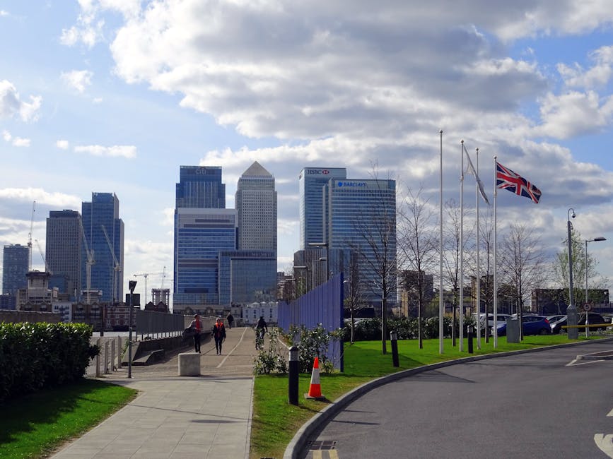 A daytime scene of a paved walkway leading towards a cluster of modern office skyscrapers with reflective glass facades in a city skyline. In the foreground, there is a small grassy area with a black metal fence, along with young, leafless trees and several flagpoles, one of which is flying a Union Jack flag. To the right, a curved driveway with parked cars and orange traffic cones is visible. The sky above is partly cloudy with patches of blue, and there are a few pedestrians walking along the pathway, some wearing high-visibility jackets. The scene is set in an urban environment that could relate to private waste collection services operating in commercial or public areas, with the surrounding landscape carefully maintained and organized for pedestrian access and vehicle movement.